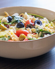  healthy orzo salad served in a bowl, made with fresh vegetables, black olives, and crumbled feta cheese close up