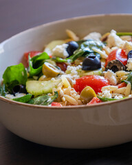  healthy orzo salad served in a bowl, made with fresh vegetables, black olives, and crumbled feta cheese close up