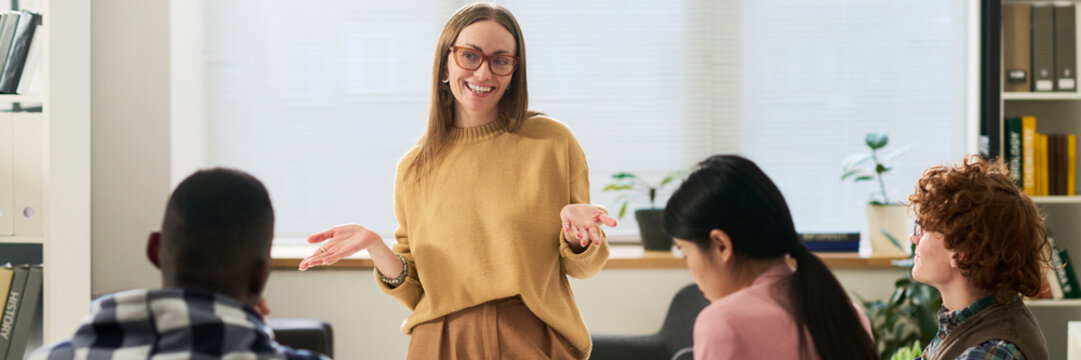 Group of diverse students engaging in studying foreign languages in bright classroom setting. Teacher standing and explaining with smiling face, surrounded by attentive students