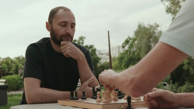 Two male friends are playing chess on a park bench on a cloudy summer day. One friend is thinking about his next move.
