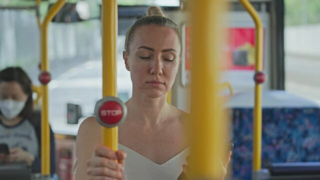 A young woman stands inside a moving city bus, holding onto a yellow handrail while using her smartphone during the ride. Urban public transport scene showing daily commute and modern mobile
