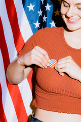 A young woman affixes a small American flag pin to her top while smiling, standing confidently...