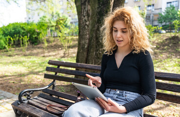Obraz premium Young girl or woman sitting in the park using a digital tablet to pass the time, surfing the internet while waiting for a friend or boyfriend to spend time together.