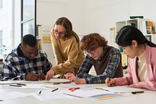 Group of diverse individuals engaging in studying foreign languages in classroom setting, with books and papers on table, and teacher assisting students during their learning process