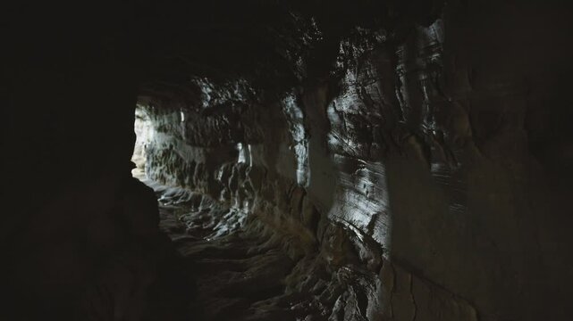 First-person view of narrow stone cave passage with visible light at the end. Mysterious underground tunnel with rough rocky walls and natural illumination.