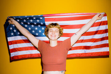 Fototapeta premium A cheerful young woman with short hair beams while holding a large American flag. The bright yellow background adds to the lively atmosphere of patriotism and joy.