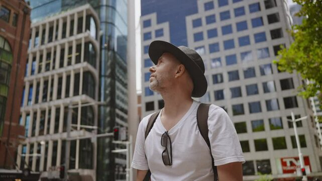 A man wearing a black hat, white t-shirt, and a backpack is standing in the middle of a modern city, looking up at the tall glass and concrete buildings surrounding him.
