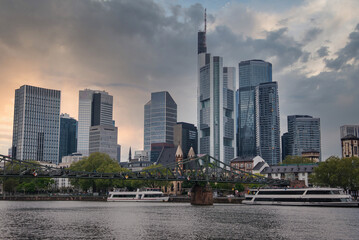 Obraz premium The skyline of Frankfurt, Germany, features modern skyscrapers like Commerzbank Tower, the Main River with boats, and the historic Eiserner Steg bridge.