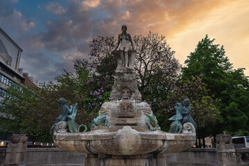 Obraz premium The Marchenbrunnen Fountain in Frankfurt features a central stone sculpture, bronze figures of children and animals, lush trees, and urban architecture.