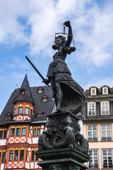 Obraz premium The Fountain of Justice statue in Frankfurt's Romerberg square with Lady Justice, surrounded by half timbered buildings under a bright blue sky.