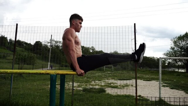 Strong man trains abdominal muscles raising legs on parallel bars at sports ground. Young shirtless sportsman exercising at nature. Sporty athlete working out at nature. Concept of sportive lifestyle