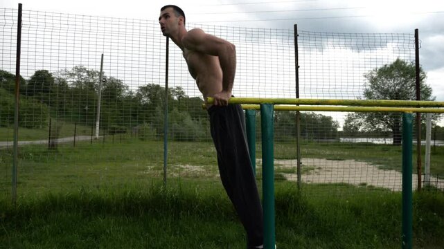 Young sportsman doing push ups on parallel bars at nature. Strong sporty man exercising at sports ground. Male shirtless athlete training hard outdoor. Concept of active sportive lifestyle. Slow mo