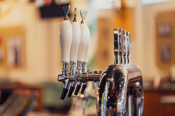 A close-up view of a polished beer tap featuring multiple handles. The shiny metal contrasts with the ceramic-white tap handles, set in a warm, inviting bar environment.