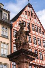 Obraz premium A detailed statue of a warrior holding a spear stands on an ornate pedestal, with a red and white half timbered building and modern architecture in Frankfurt.