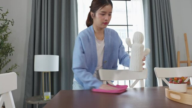 Asian young woman wearing blue sweater wipes wooden table with pink cloth, tidying up cozy living space. Home cleaning, organization, and sense of cleanliness and care for personal living environments