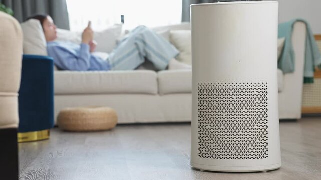 modern air purifier placed prominently in foreground of cozy living room. In the background, woman relaxes on comfortable white sofa while using smartphone, peaceful and clean home environment