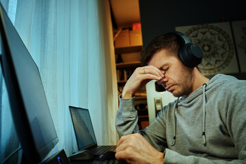 Tired man rubs eyes while working at computer. Fatigued freelancer working late at home office....