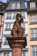 Obraz premium A detailed statue of a warrior in ornate armor holding a spear and shield, set against pastel colored European style buildings in Frankfurt, Germany.