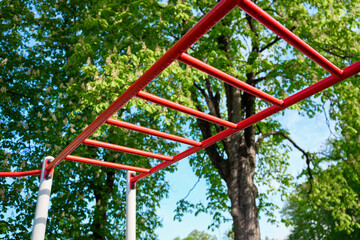 Red outdoor monkey bars in park with green trees. Fitness playground horizontal ladder for sport exercises. Concept of outdoor fitness, calisthenics training and healthy active lifestyle