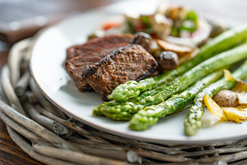 Roasted green asparagus with a beef steak, mushrooms, and crispy potato slices served on a white plate. Close-up with shallow depth of field.