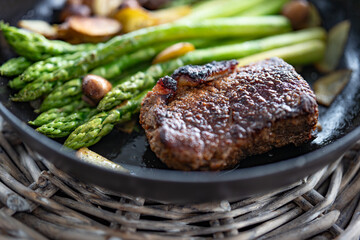 Crispy roasted beef steak with green asparagus, mushrooms, and potato slices in a cast iron pan. Close-up with shallow depth of field.