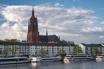 Fototapeta premium The Frankfurt Cathedral's Gothic spire rises against a cloudy sky, with the Main River, docked boats, and modern residential buildings in the foreground.