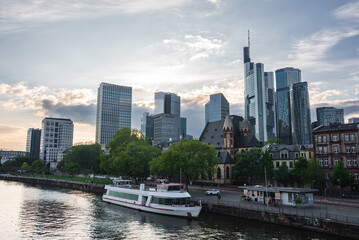 Skyline of Frankfurt, Germany, featuring modern skyscrapers like Main Tower, a historic church, the River Main, a docked boat, and green trees.