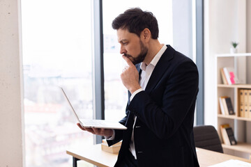 Naklejka premium Young adult Caucasian male working on laptop in office with large windows and shelves. Represents productivity, technology, and professional lifestyle.