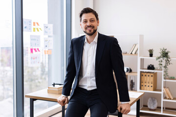 Fototapeta premium Middle-aged man wearing suit sitting on desk in well-lit modern office setting, exuding professionalism and confidence. Office surroundings include books, charts, and plants.