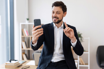 Adult Caucasian man wearing suit engaged in video call through smartphone indoors. Office in...