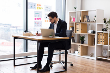 Businessman wearing formal suit working at desk, planning and making notes, in modern office with bright environment and natural light. Workspace includes organized shelving and stationery.