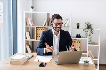 Young adult male in suit and headset smiling during online meeting, working at desk in bright office, expressing professional engagement and effective communication.