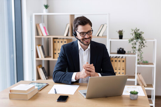 Young adult male in suit and headset smiling during online meeting, working at desk in bright office, expressing professional engagement and effective communication.