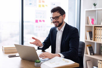 Professional adult male in suit smiling during video call in modern office setting. Displays confident gesture while laptop is seen on desk, making connection remotely.