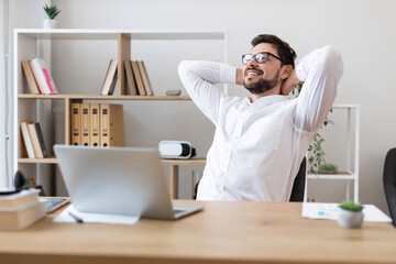 Young adult white male wearing glasses and white shirt sitting at desk smiling during office break, showcasing relaxation and positive work-life balance.