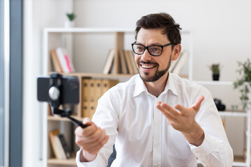 Middle-aged man recording video call with smartphone indoors expressing happiness and engagement. Dressed in white shirt, captured in bright modern office environment.