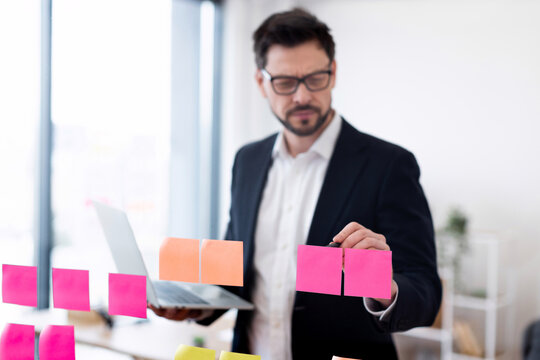 Professional adult male organizing tasks using colored sticky notes on glass. Indoors workspace setting emphasizing planning, productivity with business attire, natural light setup.