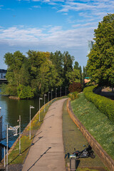 A paved riverside pathway in Frankfurt, Germany, bordered by greenery and a calm river. A motorcycle is parked near the path, with a skyscraper visible.