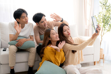 Group young asian friends sitting on sofa taking a selfie or video call using digital tablet in living room at home, cheerful and smiling while connection to social media, interaction and friendship.