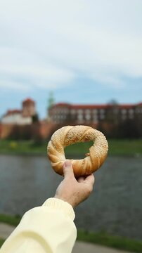 Hand holding a traditional Krakow bagel (obwarzanek) against the backdrop of the historic Wawel Castle and Vistula River. A perfect combination of local food, culture, and travel in Poland.