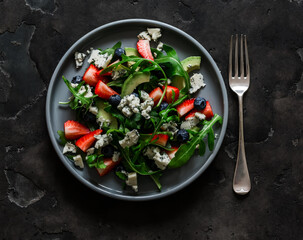 Fresh salad with avocado, strawberries, arugula, blueberries and gorgonzola on a dark background, top view