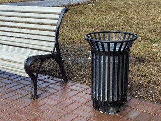 A black metal trash can in a spring park, next to a bench