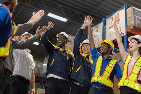 Group of warehouse workers joyfully high-fiving each other, showing teamwork and camaraderie after finishing significant task successfully. Atmosphere lively and encouraging.