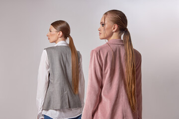 Models pose back-to-back in a studio, dressed in stylish neutral outfits with long hair pulled back. focus is on contemporary fashion and the unique textures of their clothing choices
