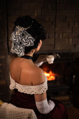 Stylisation of vintage lifestyle. Lady in noble style costume having evening tea next to the fireplace. Edwardian time hairstyle, krochet hairpeace, white gloves.