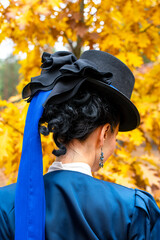 Black cylinder hat with blue ribbon and black bow (backsword view). Victorian fashion attributes, abstract, close-up. Retro lady, vintage walking outfit in blue color. Glossy deep blue satin jacket. 