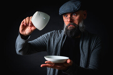 Surprised bearded man in flat cap holding an empty cup and saucer, checking it with raised eyebrows. Comical expression, studio shot with dark background.