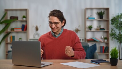 Fototapeta premium Young adult man spending time at home, man student sits at desk works on laptop, received exam results, happy expression winning gestures after passing the test.