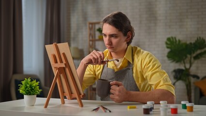 Young adult man spending time at home, man in apron sits at the table drinks tea looks at picture on portable easel, thinking and doubting, no inspiration.
