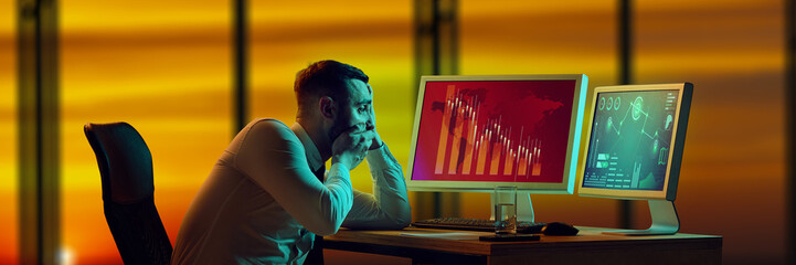 Shocked businessman staring at desk, looking at monitors showing collapsing financial charts, total...